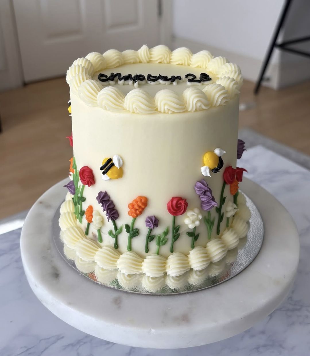 Tall cream vintage cake with shell-piped borders, “Chapter 26” written on top and hand-piped wildflowers and bees around the side (photo 2 of 3)