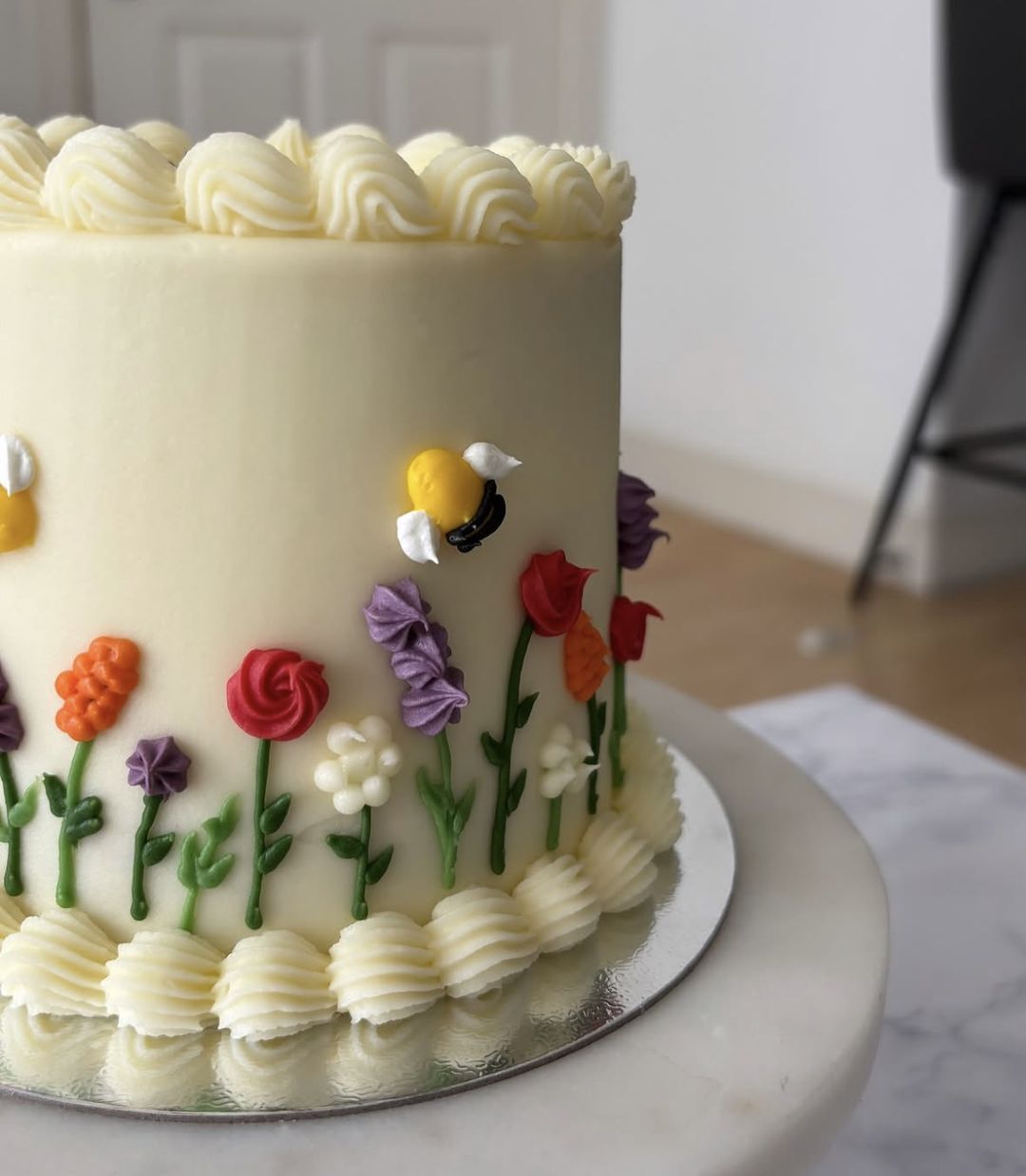 Tall cream vintage cake with shell-piped borders, “Chapter 26” written on top and hand-piped wildflowers and bees around the side (photo 3 of 3)