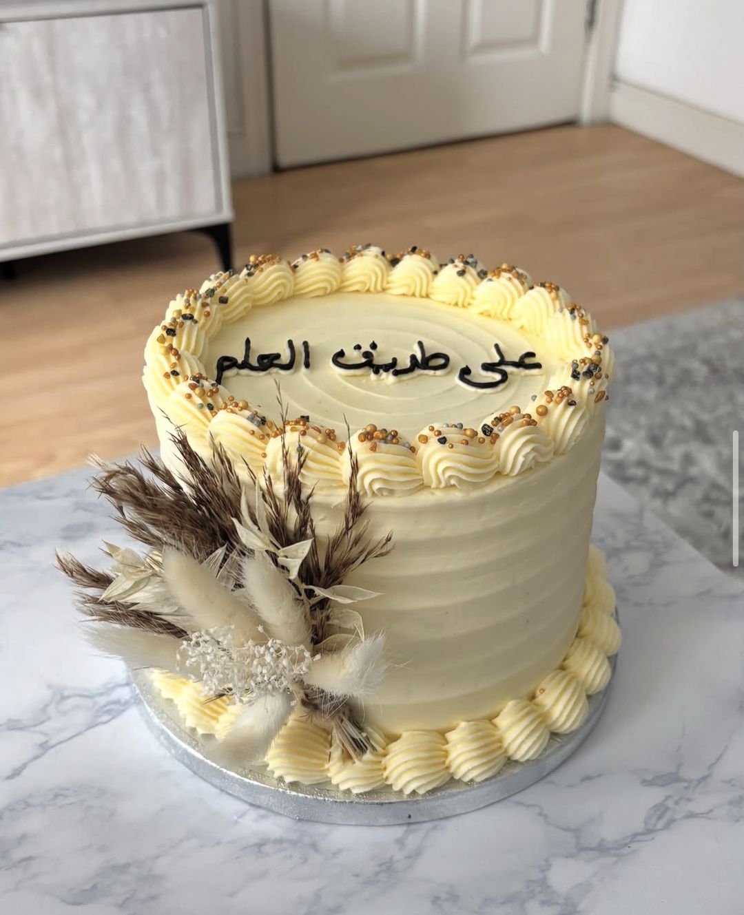 Cream cake with gold-sprinkled piping, Arabic lettering on top and dried pampas flowers on the side (photo 1 of 3)